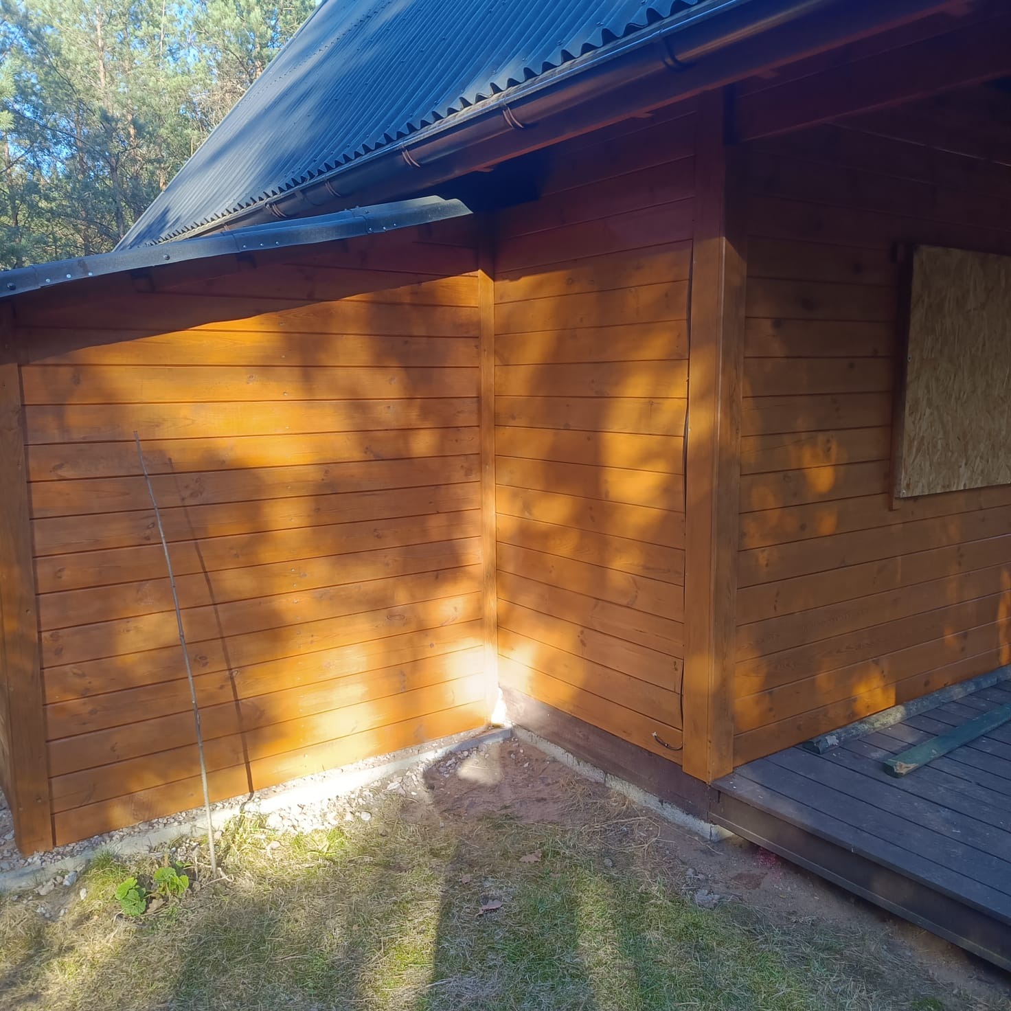 Drewniana elewacja budynku po impregnacji, widoczne deski w ciepłym odcieniu brązu, cień drzew na ścianie, fragment dachu i podmurówki.