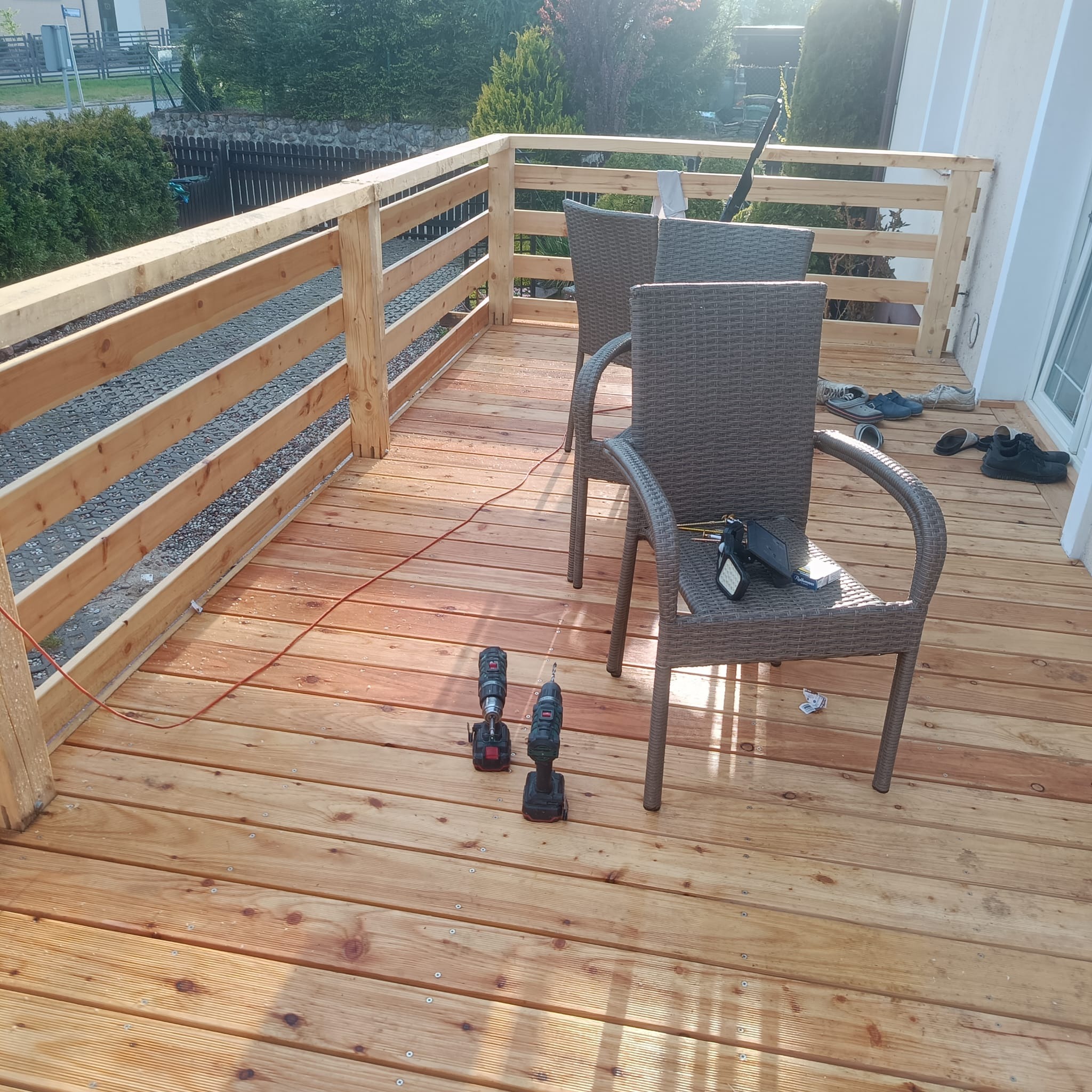 Nowa drewniana podłoga tarasu z balustradą, dwa krzesła z narzędziami i butami. Widok na zewnątrz, słoneczny dzień.