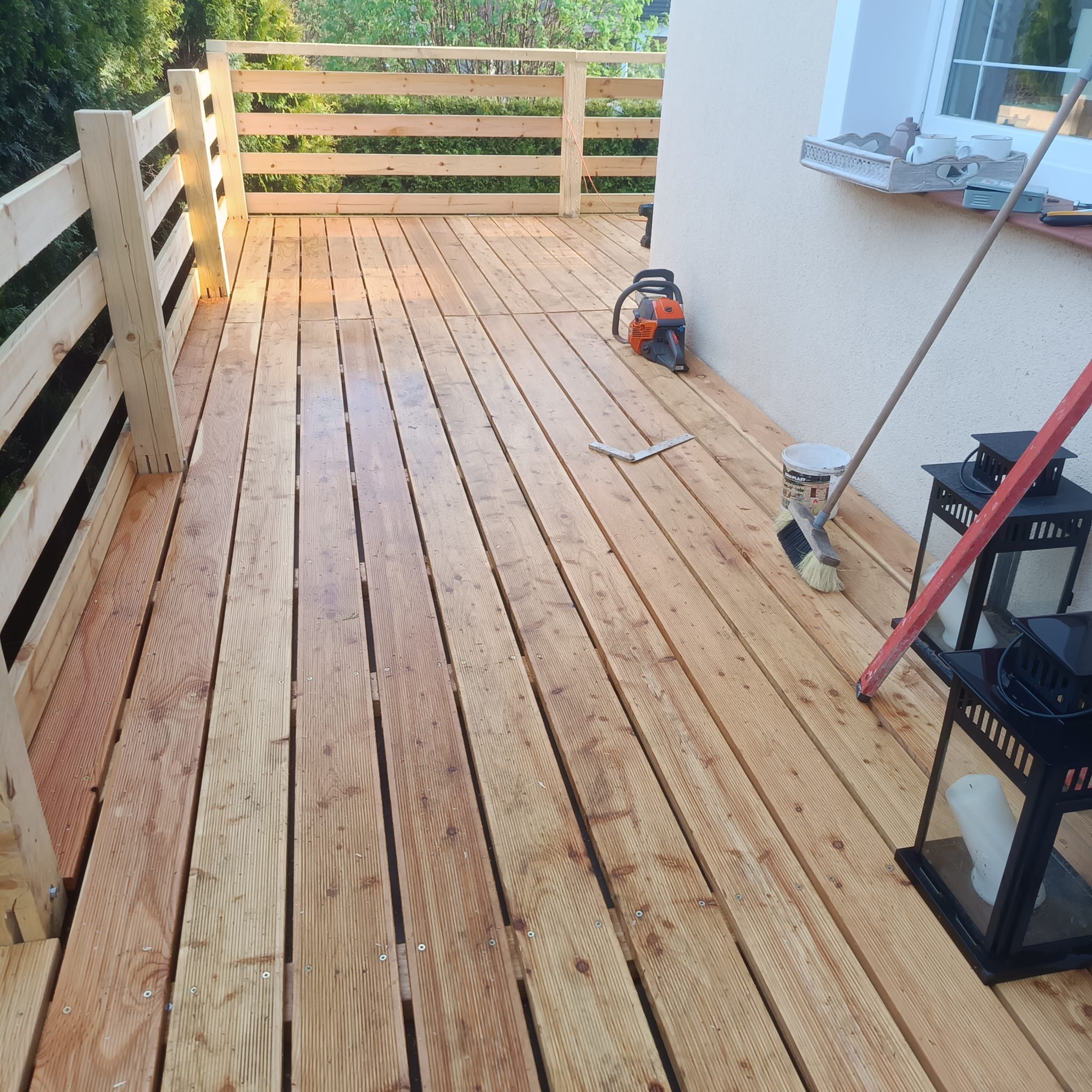 Drewniany taras z jasną balustradą, widoczne narzędzia i akcesoria do czyszczenia, sugerujące świeżo zakończone prace wykończeniowe.