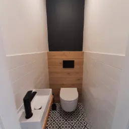 Nowoczesna, wąska łazienka w Ząbkach po remoncie: białe płytki imitujące drewno na ścianach do połowy wysokości, nad nimi białe ściany, drewniana boazeria za toaletą, umywalka nablatowa z czarną...