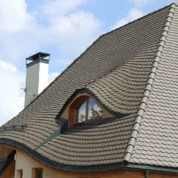 Dach pokryty łuską ceramiczną w odcieniach szarości z oknem dachowym w kształcie oka, widoczny komin i rynna w kolorze ciemnej zieleni.