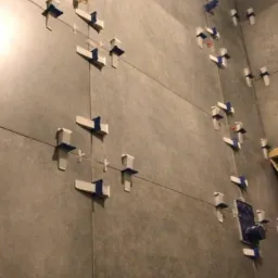 Szare płytki wielkoformatowe na ścianie łazienki, w trakcie montażu z użyciem białych klinów i niebieskiej taśmy, widoczny fragment instalacji wodnej.