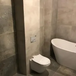 Nowoczesna łazienka z szarymi płytkami imitującymi beton, wisząca toaleta z chromowanym przyciskiem spłukującym i wolnostojąca wanna.