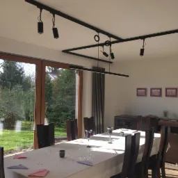 Jadalnia z nowoczesnym oświetleniem szynowym i lampą wiszącą LED, widok na ogród przez duże okno, stół nakryty do uroczystości.