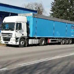 Biała ciężarówka DAF z niebieską naczepą marki Knapen Trailers zaparkowana na poboczu drogi, z budynkiem przemysłowym w tle.