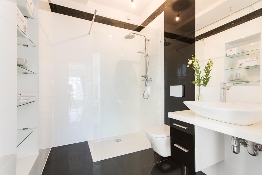 Nowoczesna łazienka w bieli i czerni z kabiną prysznicową typu walk-in, umywalką nablatową i minimalistycznymi półkami szklanymi.