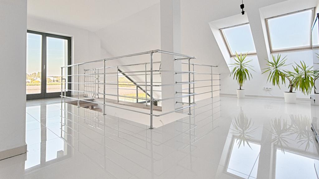 Jasne, nowoczesne wnętrze w Zebrzydowicach z błyszczącą białą podłogą, metalową balustradą, dużymi oknami dachowymi i roślinami w doniczkach.