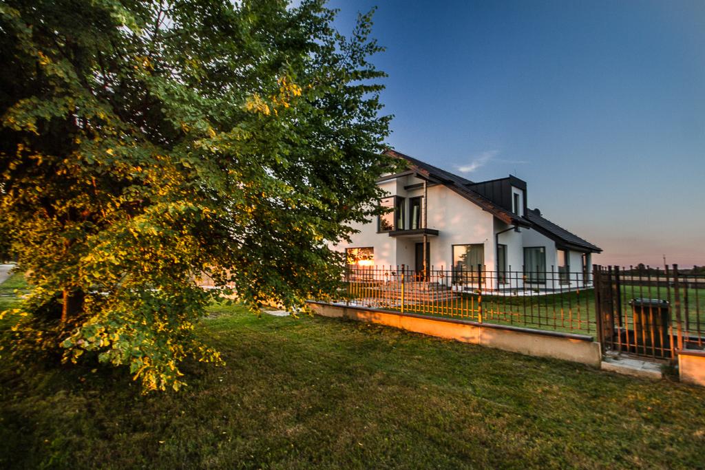 Nowoczesny dom jednorodzinny z białymi ścianami i czarnym dachem, widziany z perspektywy ogrodu oświetlonego zachodzącym słońcem, z drzewem w lewym rogu kadru.