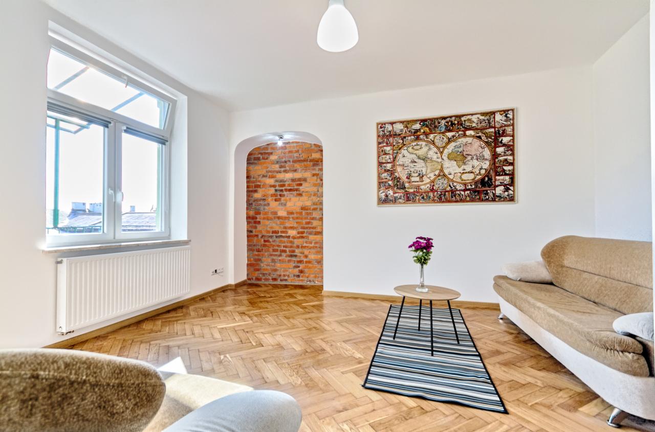 Jasne wnętrze mieszkania z parkietem w jodełkę, ceglaną wnęką, sofą, stolikiem z kwiatami i mapą świata na ścianie.