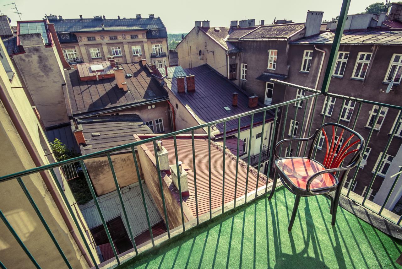 Widok z balkonu na stare kamienice, fotel z czerwonym oparciem na sztucznej zielonej trawie, zielona balustrada, perspektywa z góry.