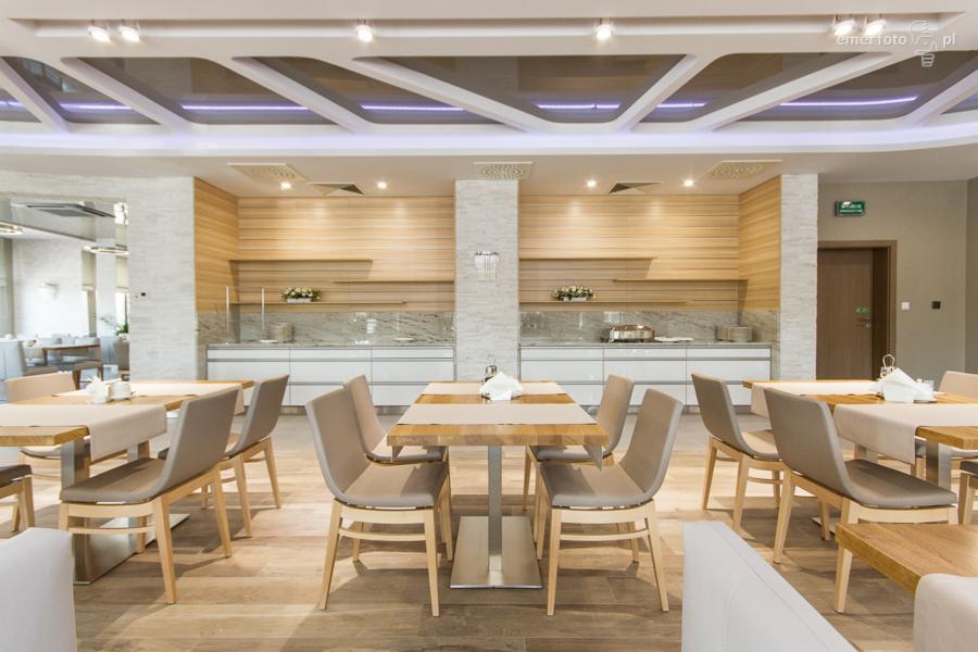 Jasne, przestronne wnętrze restauracji hotelowej z drewnianymi stołami i krzesłami, minimalistyczną kuchnią w tle oraz dekoracyjnym sufitem z oświetleniem LED.