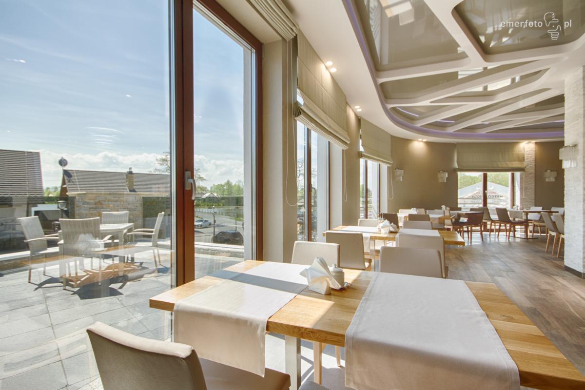 Jasne wnętrze restauracji z drewnianymi stołami nakrytymi białymi obrusami, widok na taras z krzesłami i zabudowę mieszkalną przez duże okna.