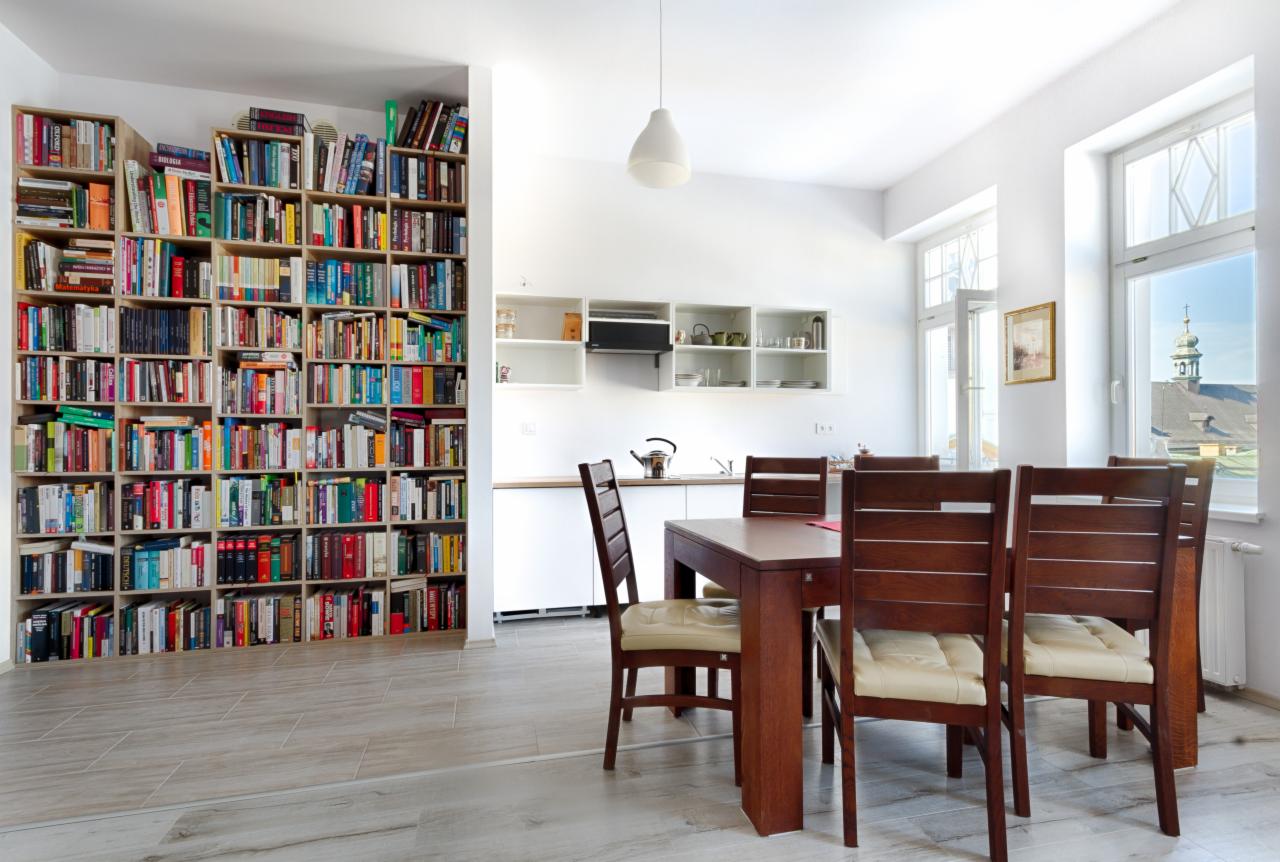 Jasne wnętrze jadalni z dużym regałem na książki, drewnianym stołem i krzesłami z kremowymi poduszkami, białą kuchnią w tle i widokiem na wieżę kościoła przez okno.