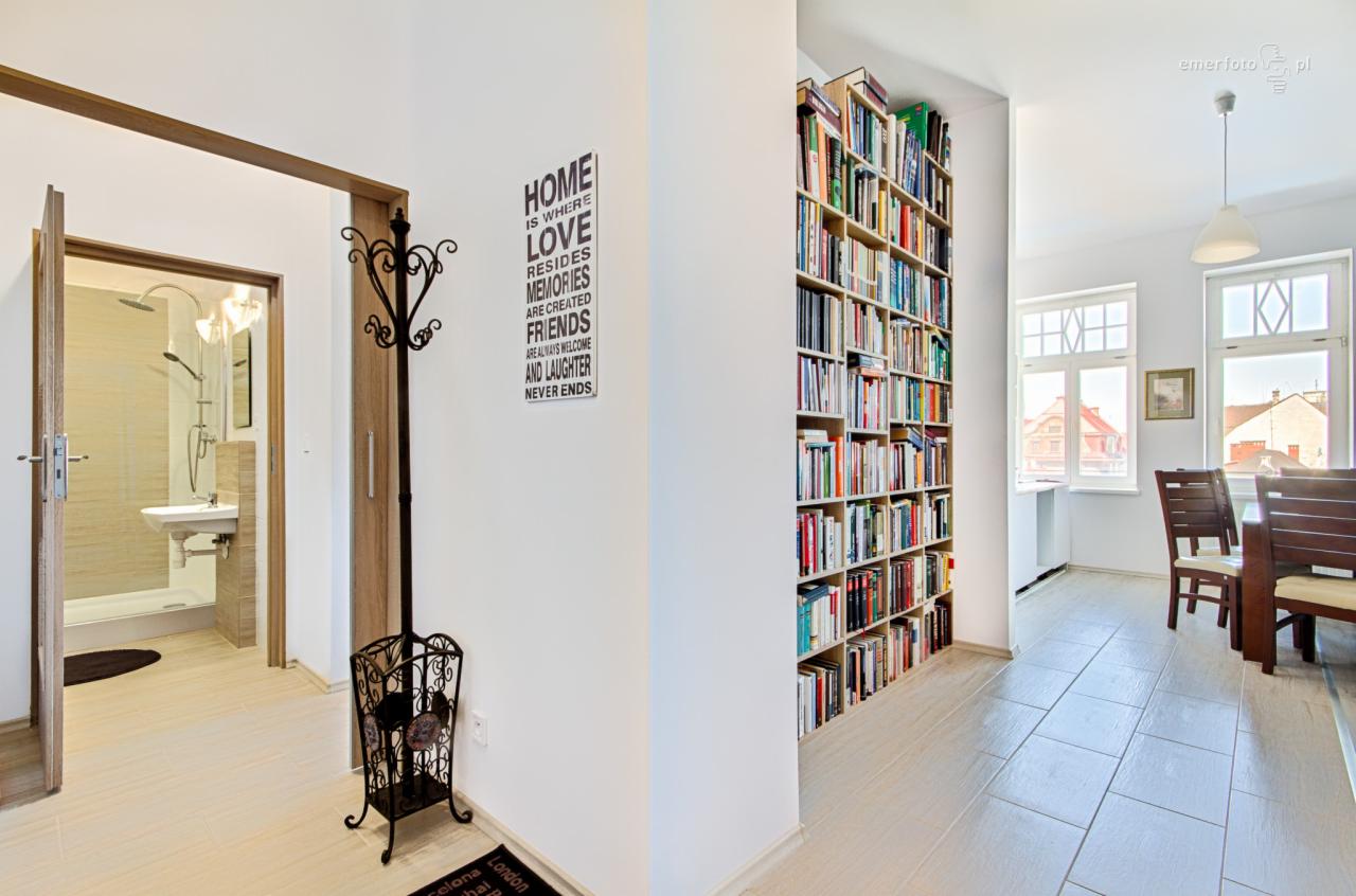 Jasne wnętrze mieszkania z widokiem na łazienkę, wysoką biblioteczkę i jadalnię, dekoracyjny wieszak na ubrania przy ścianie, napis HOME IS WHERE LOVE na ścianie.