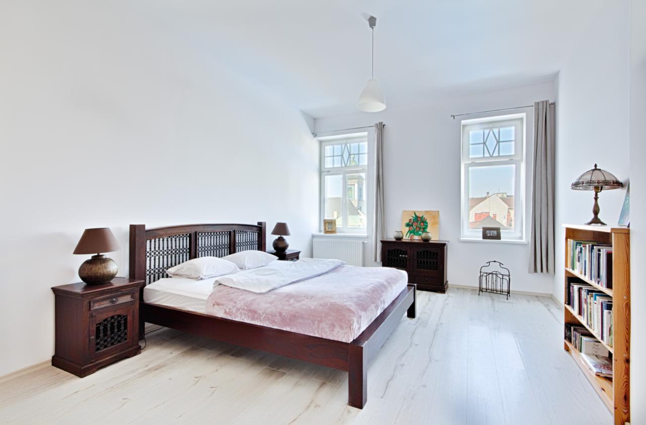 Jasna sypialnia z drewnianym łóżkiem z ażurowym zagłówkiem, nocnymi szafkami z lampkami, białymi ścianami i podłogą, oraz szafką z obrazem przy oknie.