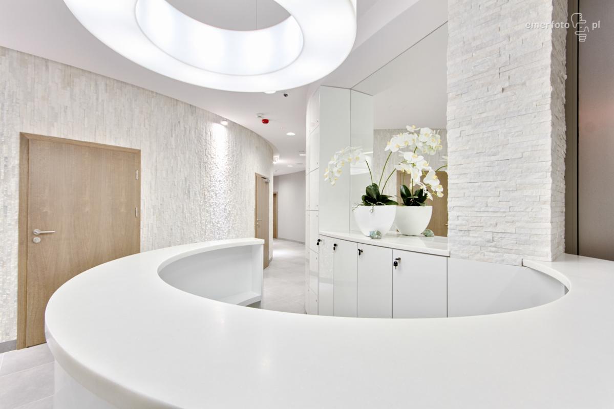 Jasne, nowoczesne wnętrze: zakrzywiona lada recepcyjna z białego konglomeratu, białe szafki z orchideami w doniczkach, ściana z białej cegły dekoracyjnej, okrągła lampa sufitowa.