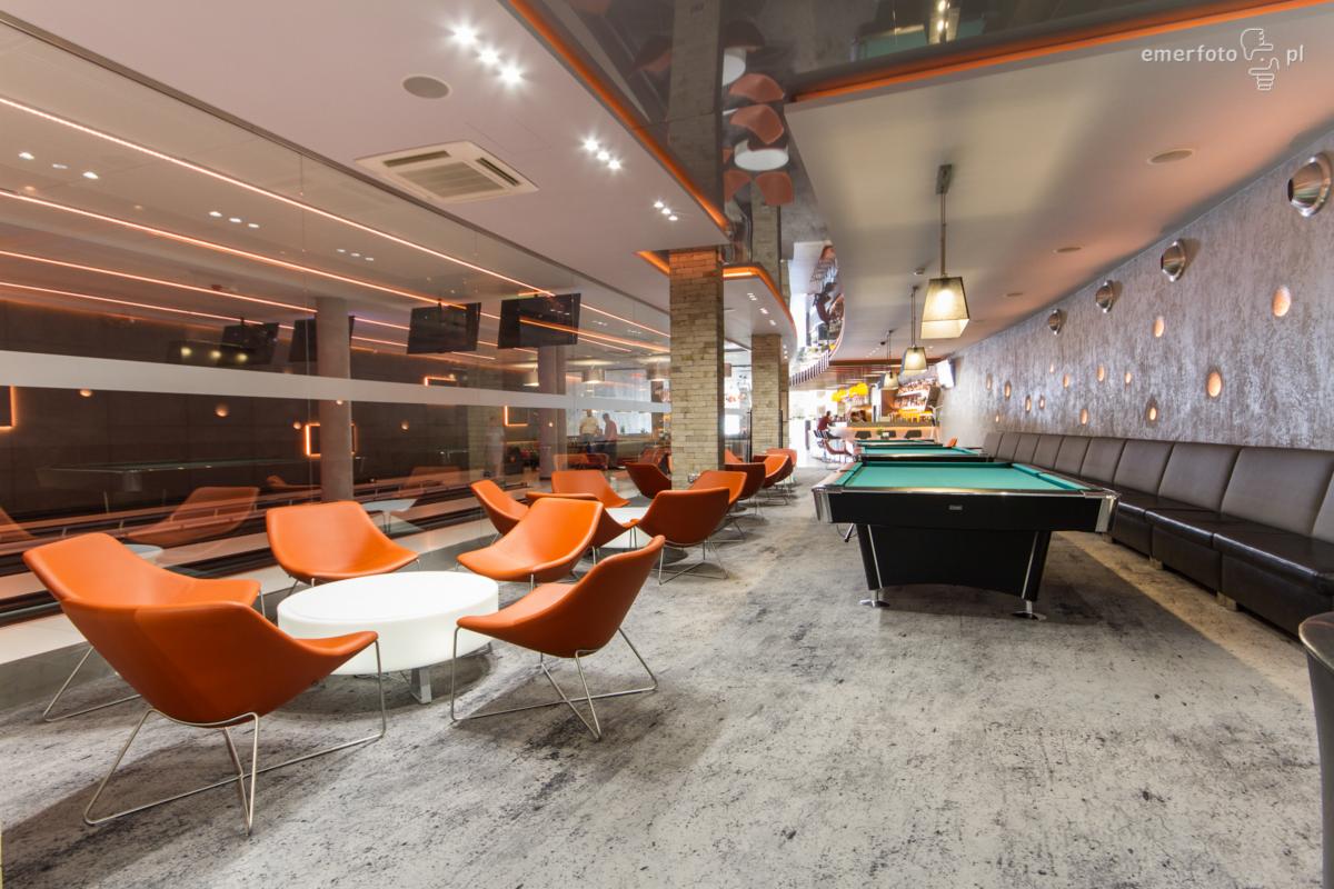 Nowoczesne wnętrze lokalu rozrywkowego z pomarańczowymi akcentami: krzesła, oświetlenie, stoliki, bilard, parkiet i bar w tle.