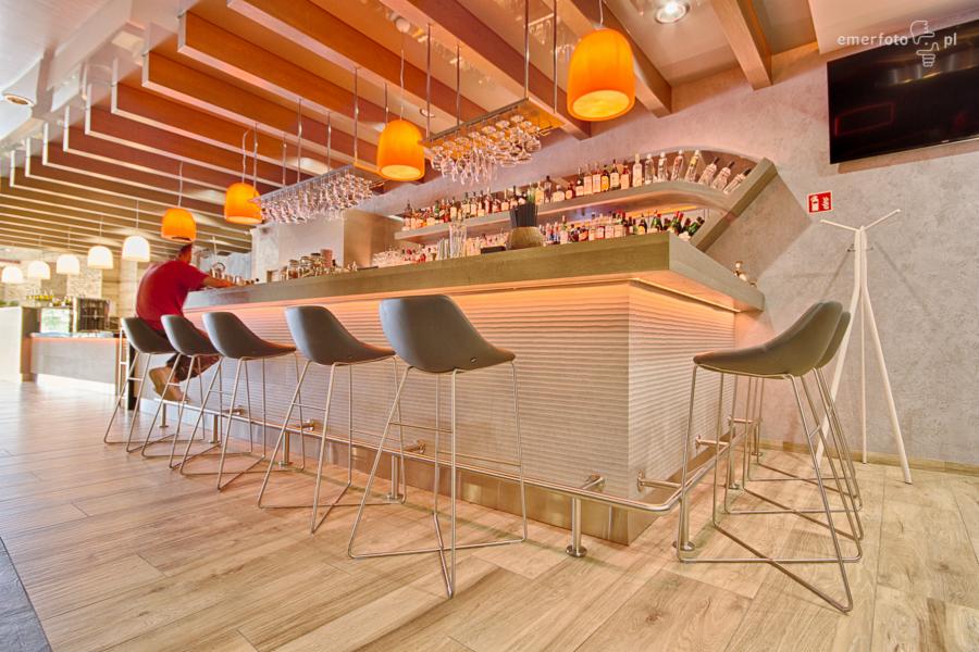 Nowoczesny bar z podświetlanym frontem, rzędem hokerów i półkami z alkoholem, oświetlony pomarańczowymi lampami wiszącymi.