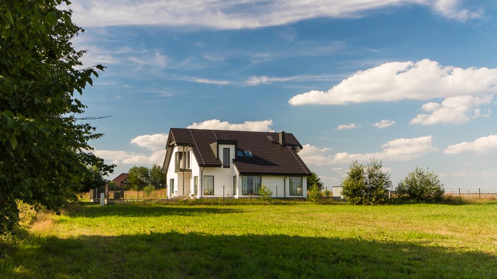 Nowoczesny, biały dom jednorodzinny z ciemnym dachem, widoczny z perspektywy trawnika w słoneczny dzień z błękitnym niebem i chmurami.