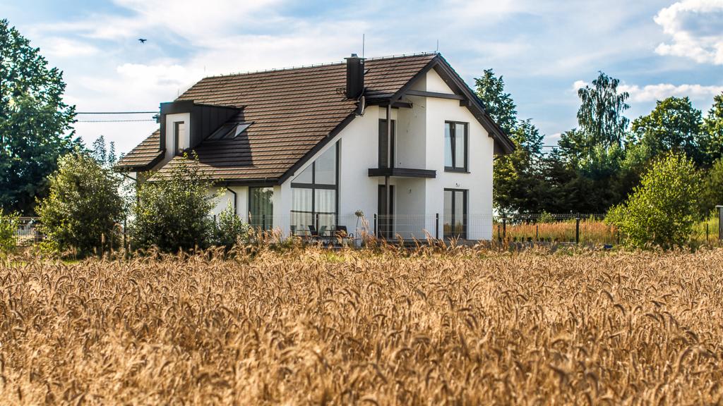 Nowoczesny, biały dom z brązowym dachem w otoczeniu pola zboża, widok z perspektywy pola, błękitne niebo z chmurami w tle.