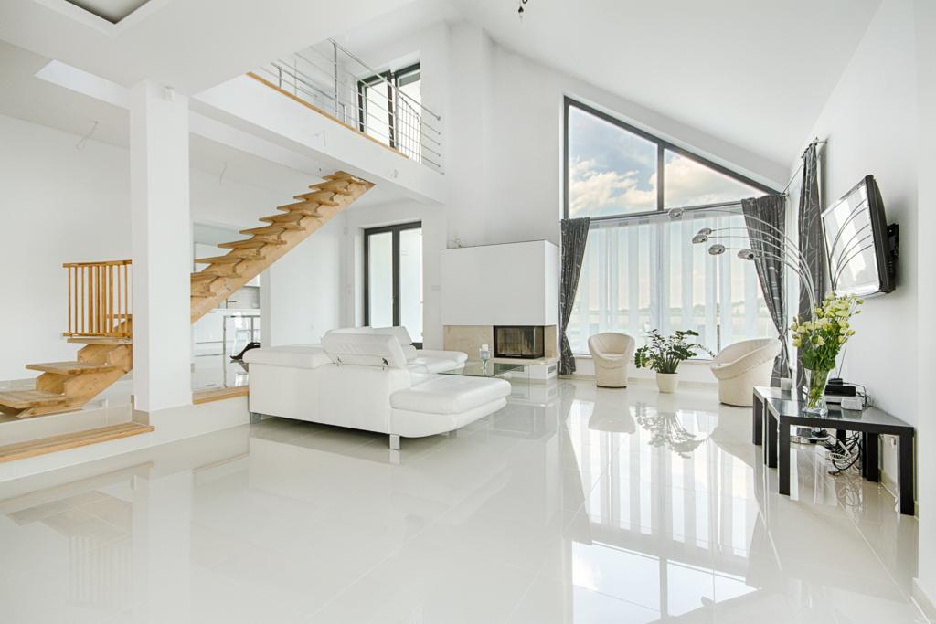 Jasne wnętrze domu z drewnianymi schodami, białymi ścianami, błyszczącą podłogą i wysokim oknem trójkątnym, z widocznym salonem z białym narożnikiem i minimalistycznym kominkiem.