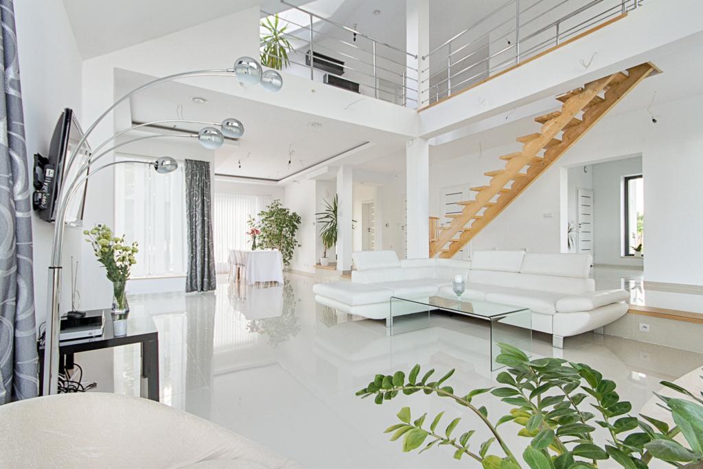 Jasne, przestronne wnętrze domu z białymi ścianami, błyszczącą podłogą, drewnianymi schodami, nowoczesną lampą i roślinami doniczkowymi.