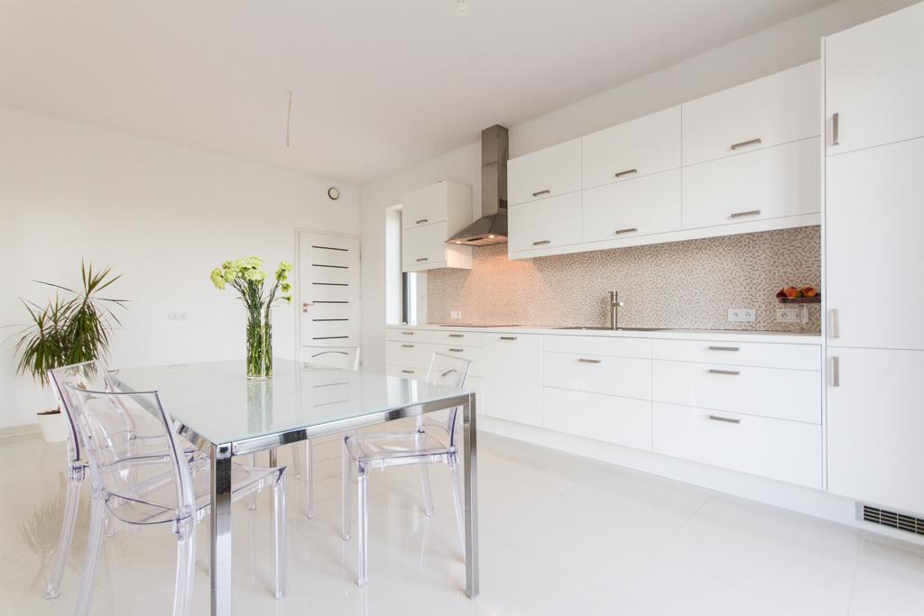 Jasna, minimalistyczna kuchnia z białymi szafkami, szklanym stołem i przezroczystymi krzesłami, uzupełniona kwiatami w wazonie i rośliną doniczkową, z mozaiką nad blatem.