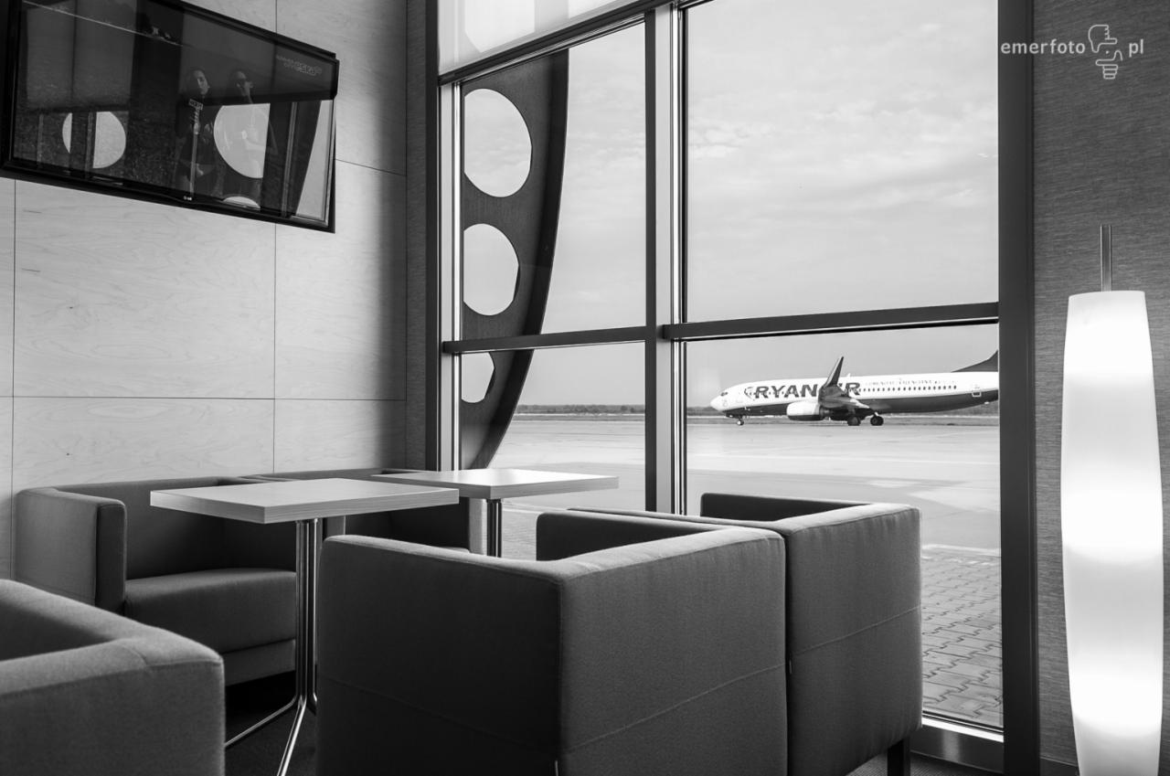 Czarno-białe zdjęcie wnętrza poczekalni z widokiem na płytę lotniska i startujący samolot Ryanair, minimalistyczny design, stoliki i fotele w odcieniach szarości, nowoczesne oświetlenie, telewizor...
