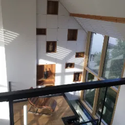 Wysoka zabudowa ścienna z białymi frontami i otwartymi półkami z jasnego drewna, widok z góry na skórzany leżak i szklany fragment podłogi przy oknie.