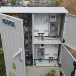 Otwarta, biała szafka elektryczna z widocznymi bezpiecznikami, przewodami i elementami instalacji, umieszczona na zewnątrz w pobliżu wykopu ziemnego.