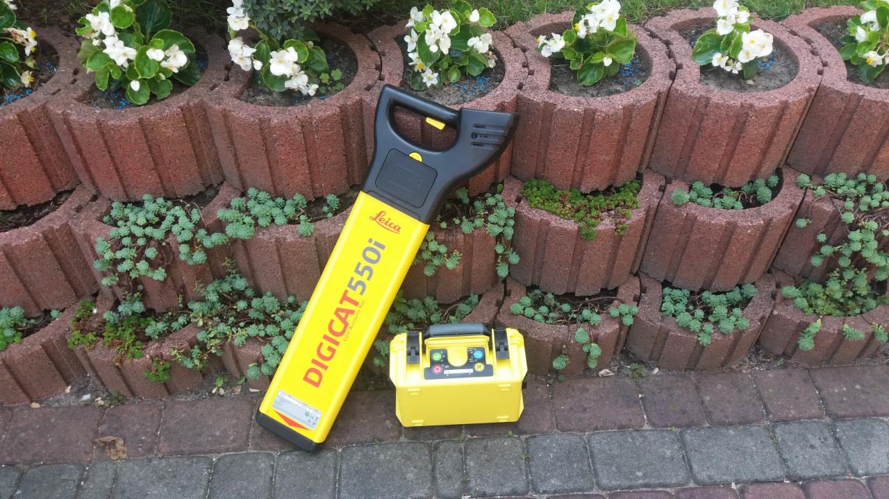 Żółty lokalizator podziemnych instalacji Leica DIGICAT 550i z walizką na kostce brukowej przy murku z kwiatami.