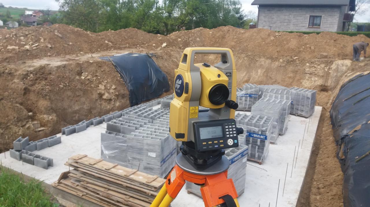 Żółty niwelator elektroniczny na trójnogu, ustawiony na fundamencie budynku w trakcie budowy, z widocznymi bloczkami fundamentowymi Bruk-Bet w folii, wykopem i ziemią w tle.