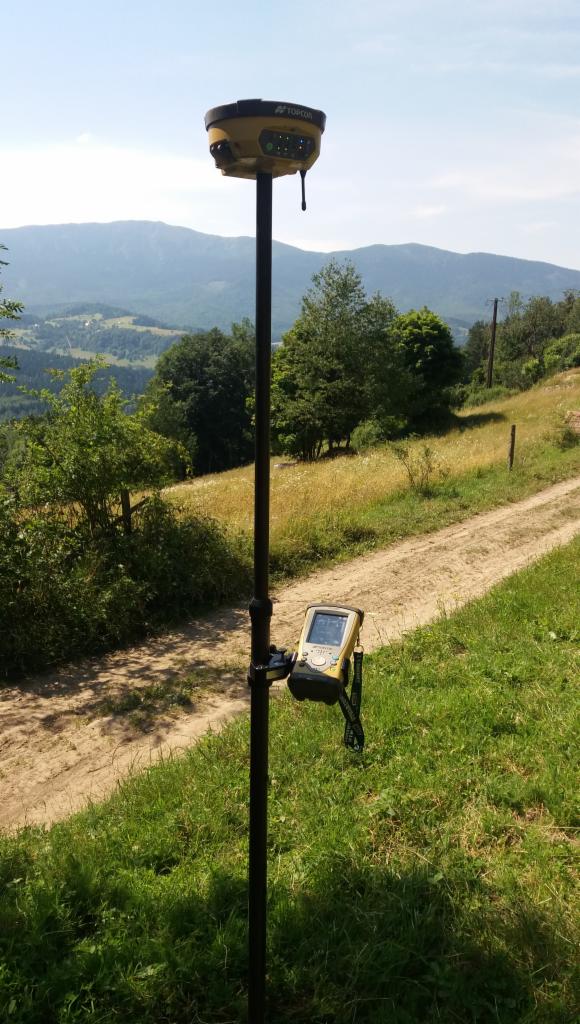 Żółty odbiornik GNSS Topcon na czarnym statywie, z kontrolerem przymocowanym do statywu, na tle górskiego krajobrazu i polnej drogi.