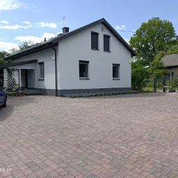 Biały dom jednorodzinny z ciemnymi ramami okiennymi i brukowanym podjazdem, błękitny samochód zaparkowany z boku.