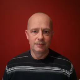 Portret mężczyzny z wąsami na tle czerwonej ściany, ubrany w sweter w paski.