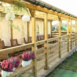 Drewniany zadaszony taras z balustradą, wyposażony w krzesła i stoliki, z wiszącymi dekoracjami i doniczkami z wrzosami, na tle zieleni i placu zabaw.