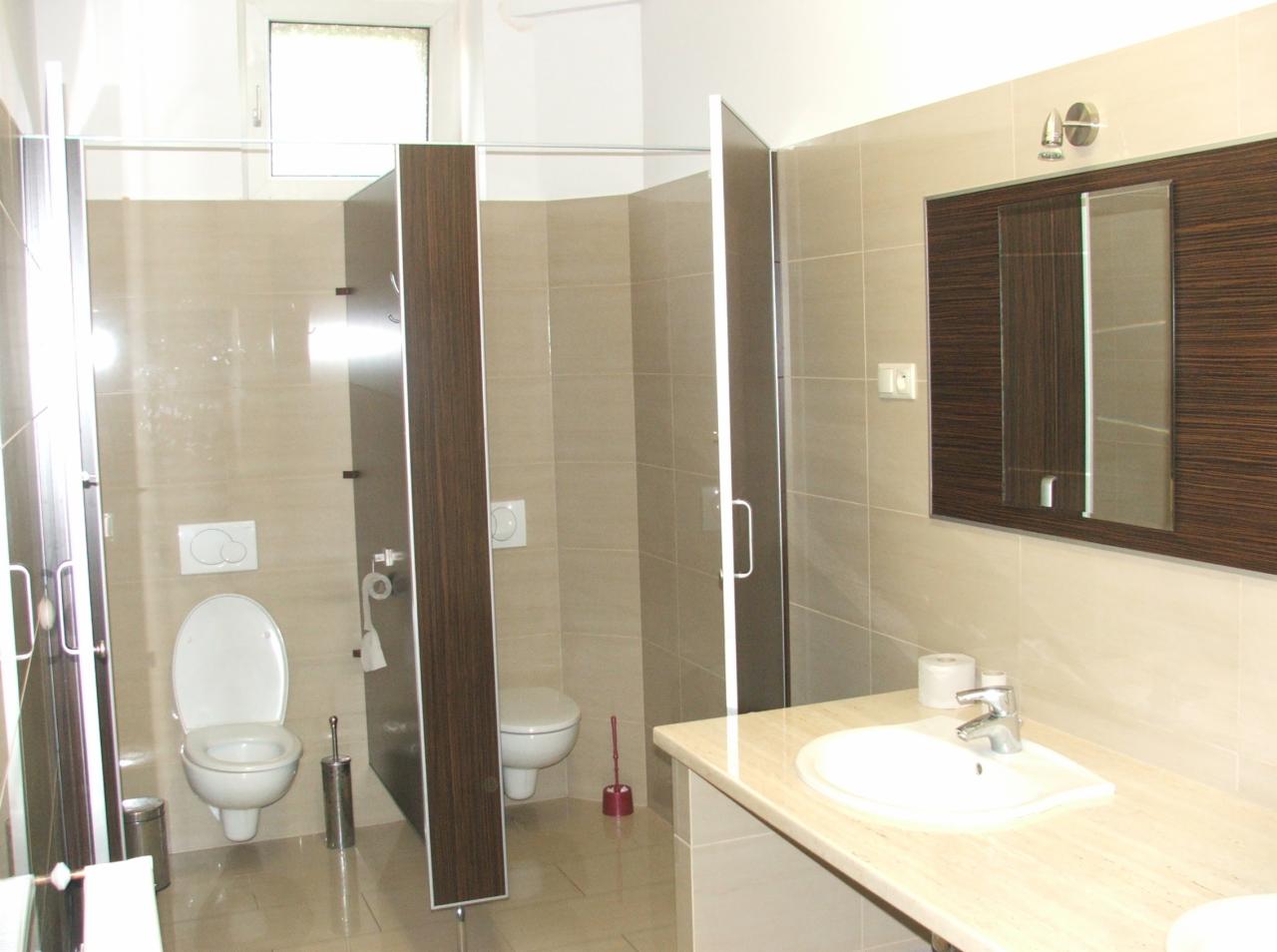 Publiczna toaleta z kabinami oddzielonymi ściankami z ciemnego drewna, jasne płytki na ścianach i podłodze, umywalka z blatem imitującym marmur, lustro w ciemnej ramie.