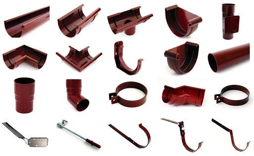 Zestaw elementów systemu odprowadzania wody deszczowej w kolorze ceglastym, prezentujący różne kształtki, rury, uchwyty i akcesoria na białym tle.