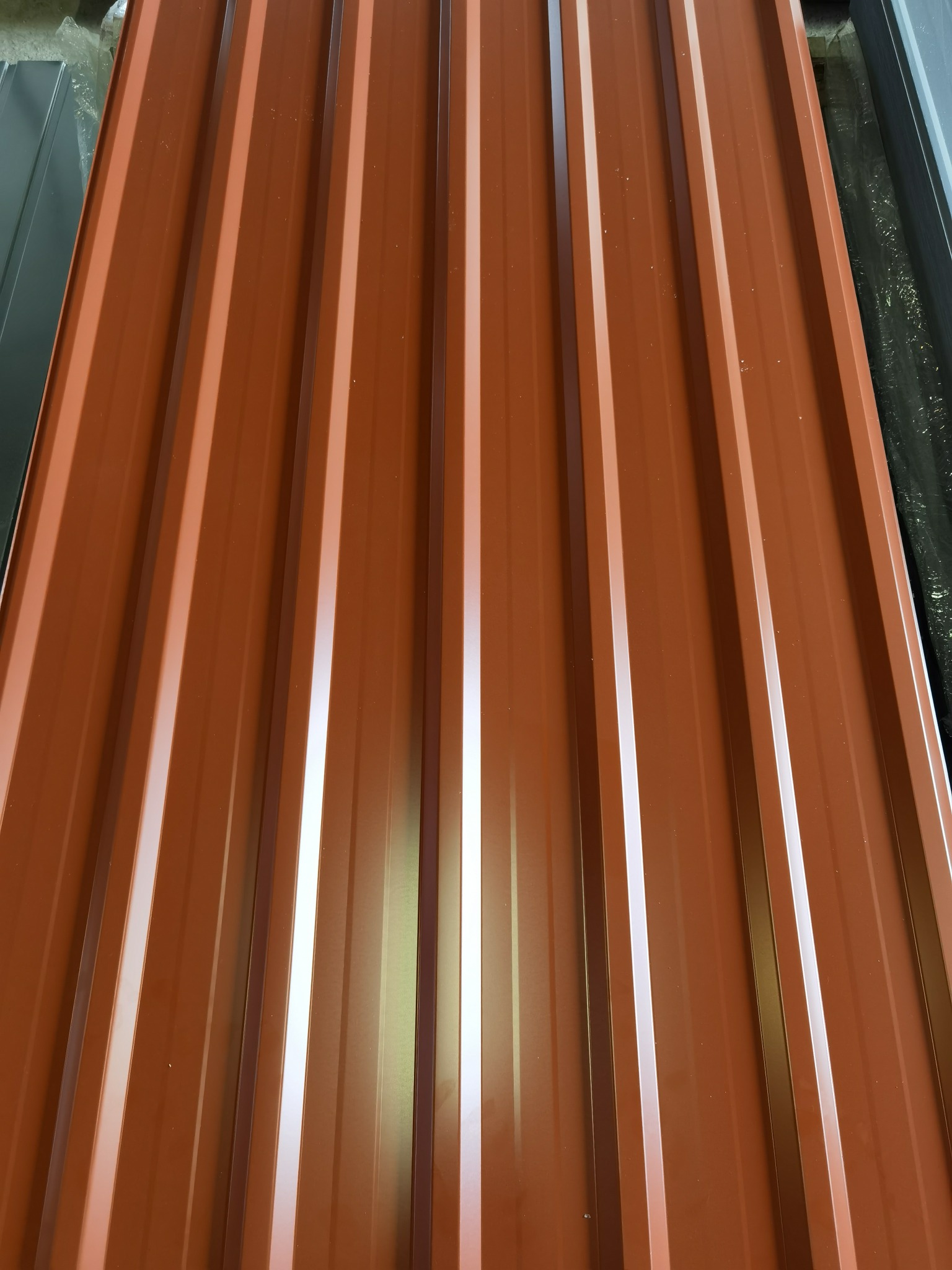 Brązowy arkusz blachy trapezowej z widocznymi przetłoczeniami, ułożony na stosie z innymi materiałami budowlanymi.