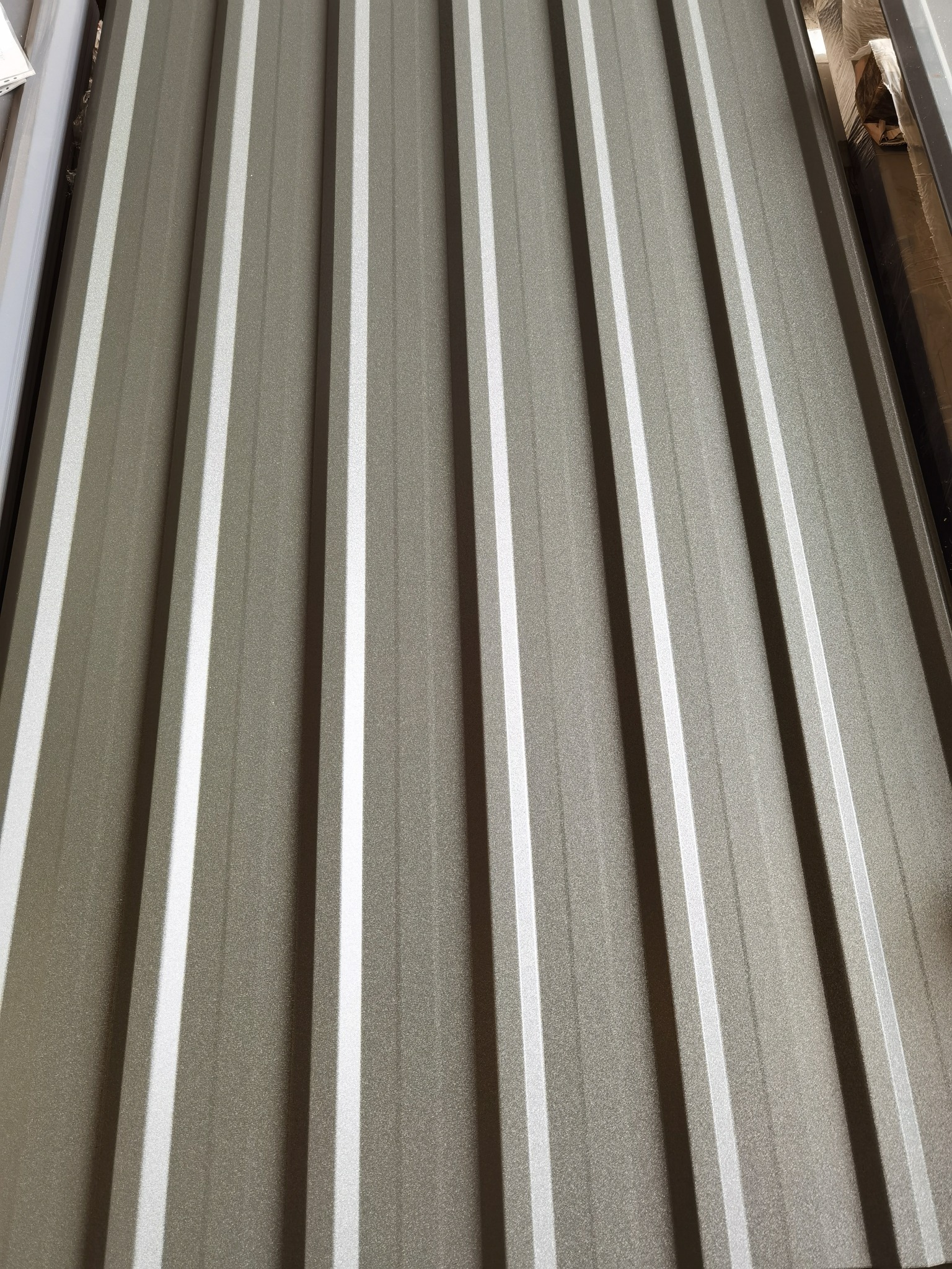 Szara blacha profilowana z wyraźnymi, pionowymi przetłoczeniami, ułożona pod kątem, z widocznym delikatnym połyskiem i fakturą.