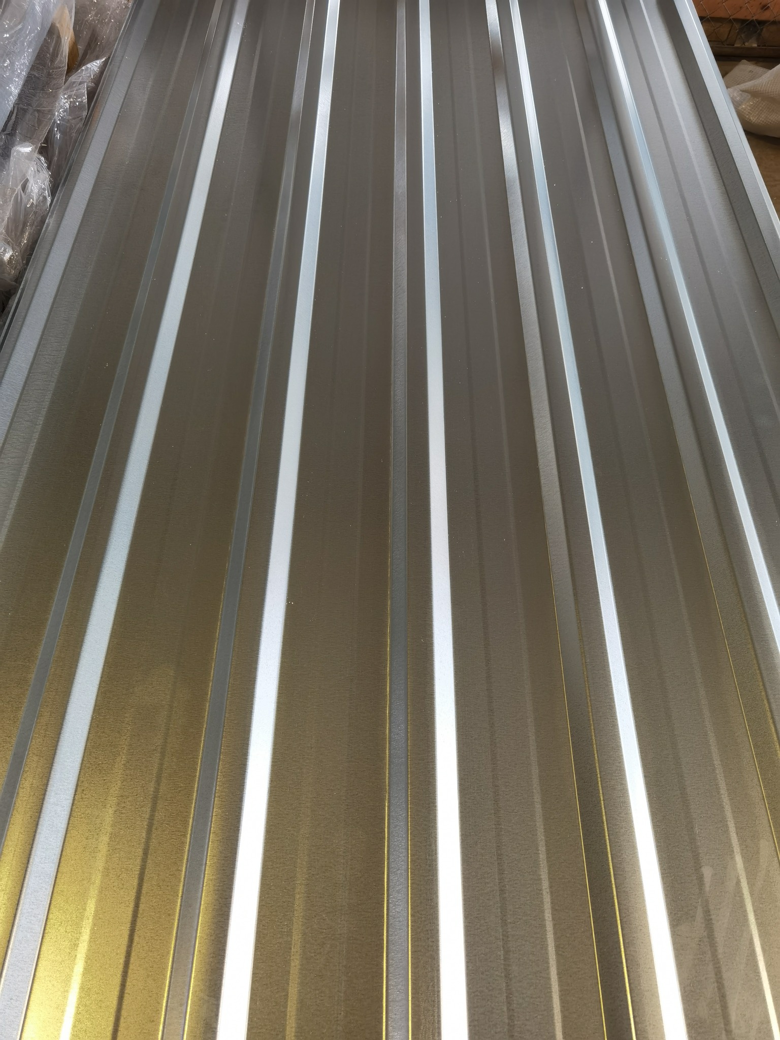 Arkusz blachy trapezowej o srebrzysto-złotym odcieniu, z widocznymi przetłoczeniami i połyskiem, ułożony pod kątem, z fragmentem folii ochronnej w lewym górnym rogu.