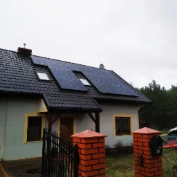 Dom jednorodzinny z zainstalowanymi panelami fotowoltaicznymi na dachu pokrytym czarną dachówką, widoczne okna dachowe i ogrodzenie z ceglanymi słupkami.