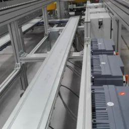 Widok na fragment hali produkcyjnej z systemem transportu wewnętrznego, widoczne szyny i szafy sterownicze z przewodami elektrycznymi.