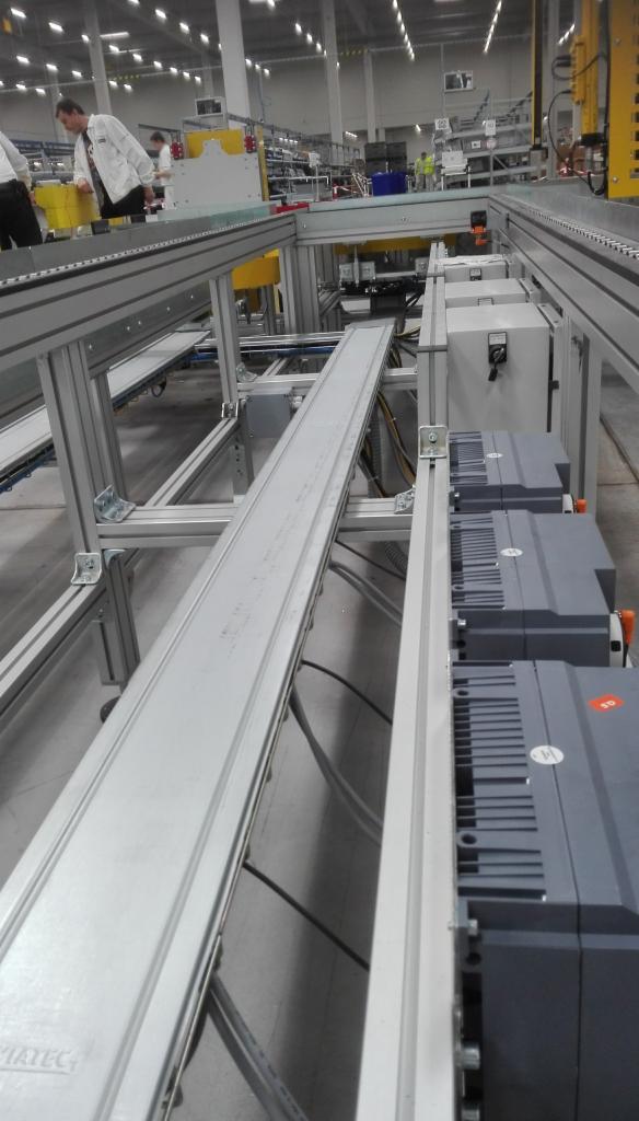 Widok na fragment hali produkcyjnej z systemem transportu wewnętrznego, widoczne szyny i szafy sterownicze z przewodami elektrycznymi.