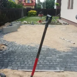 Układanie szarej kostki brukowej na podsypce piaskowej, widoczna ubijarka, łopata z czerwoną rączką, poziomica i pracownik podczas realizacji.