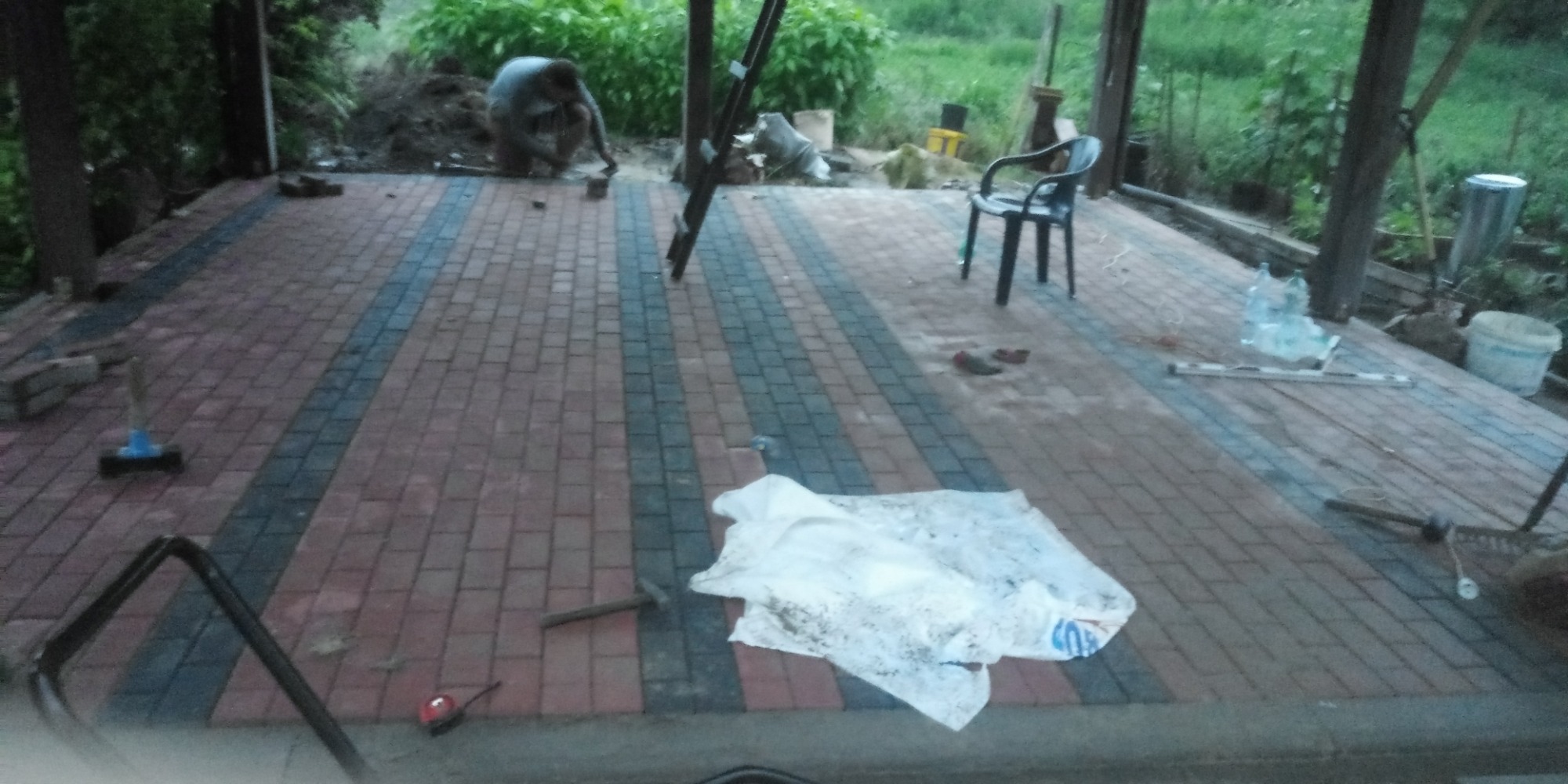 Częściowo ułożona nawierzchnia z kostki brukowej w odcieniach czerwieni i szarości na zadaszonym tarasie, z widocznym pracownikiem w tle oraz narzędziami i materiałami budowlanymi porozrzucanymi...