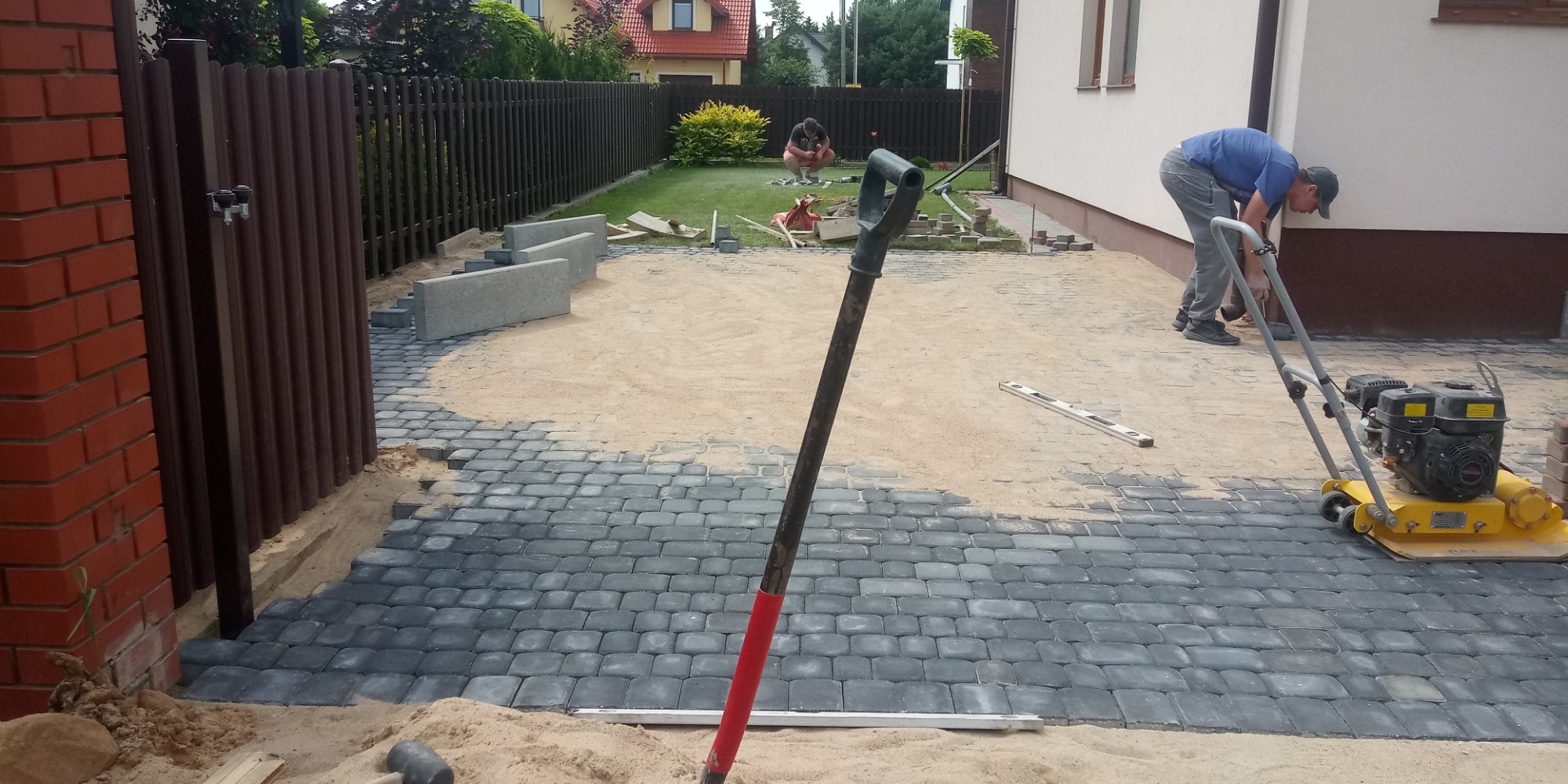Układanie szarej kostki brukowej na podsypce piaskowej, widoczna ubijarka, łopata z czerwoną rączką, poziomica i pracownik podczas realizacji.