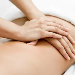 Dłonie masażysty uciskają plecy klienta leżącego na białym prześcieradle, widoczny fragment ciała i ramię masażysty.