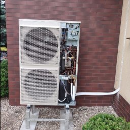 PC Eko Instalacje Przemysław Chmiel - Zewnętrzna jednostka klimatyzacji typu split, z otwartą obudową ukazującą wewnętrzne komponenty, zamontowana na betonowych podstawach przed ceglaną ścianą budynku, z widocznymi przewodami...