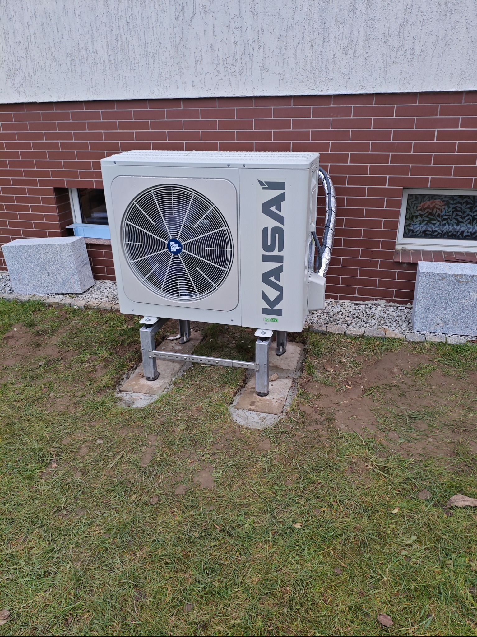 Zewnętrzna jednostka klimatyzacji Kaisai zamontowana na metalowej konstrukcji przy ścianie budynku z cegły, widoczne okno z dekoracyjną folią i kostki granitowe.